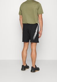 Camiseta ajustada de color verde oliva para hombre y pantalones cortos negros con un gráfico blanco de "UNDER ARMOUR" a lo largo del lateral; diseño sencillo, tejido ligero.