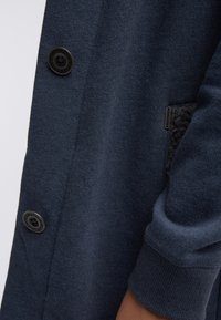 Mørkeblå cardigan med store knapper og tekstureret lomme, båret på armen med manchet synlig.