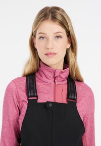 Svarta overaller med justerbara svarta remmar över en åtsittande rosa zip-up topp. Toppens högkrage och det texturerade tygmönstret är framträdande.