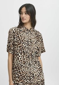 Chemise à manches courtes à motif léopard avec col classique, boutons noirs et une texture de tissu lisse et léger dans des tons beiges et marron.