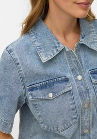 Lyseblå denimskjorte med knappestolpe foran, stor krage og to brystlommer. Har sølvfarget detaljer og en lett falmet tekstur.