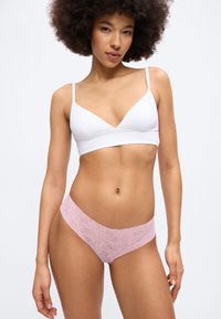 Γυναίκα με σγουρά μαλλιά, φοράει λευκό μπουστάκι και ροζ δαντελένιο εσώρουχο, μπροστά σε απλό φωτεινό φόντο.