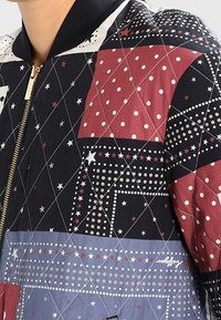 Veste bomber matelassée avec un design patchwork dans des couleurs rouge, bleu marine et blanc, avec un mélange de motifs d'étoiles et de points. Fermeture zippée dorée.