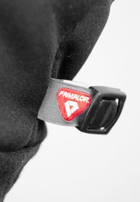 Primaloft mærketik med en rød trekantet logo på struktureret grå stof, fastgjort til en sort tekstiloverflade med en sort plastklips.