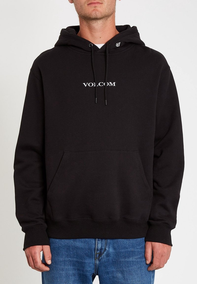 Homme portant un sweat à capuche Volcom noir avec poche frontale et jean bleu, debout sur un fond clair uni.