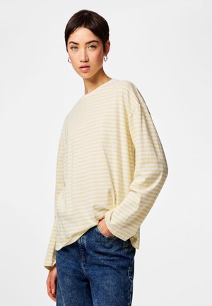 Mladá osoba s krátkými tmavými vlasy, nosící stříbrné kruhové náušnice, žlutobíle pruhované tričko s dlouhým rukávem a modré džíny, stojící uvolněně.