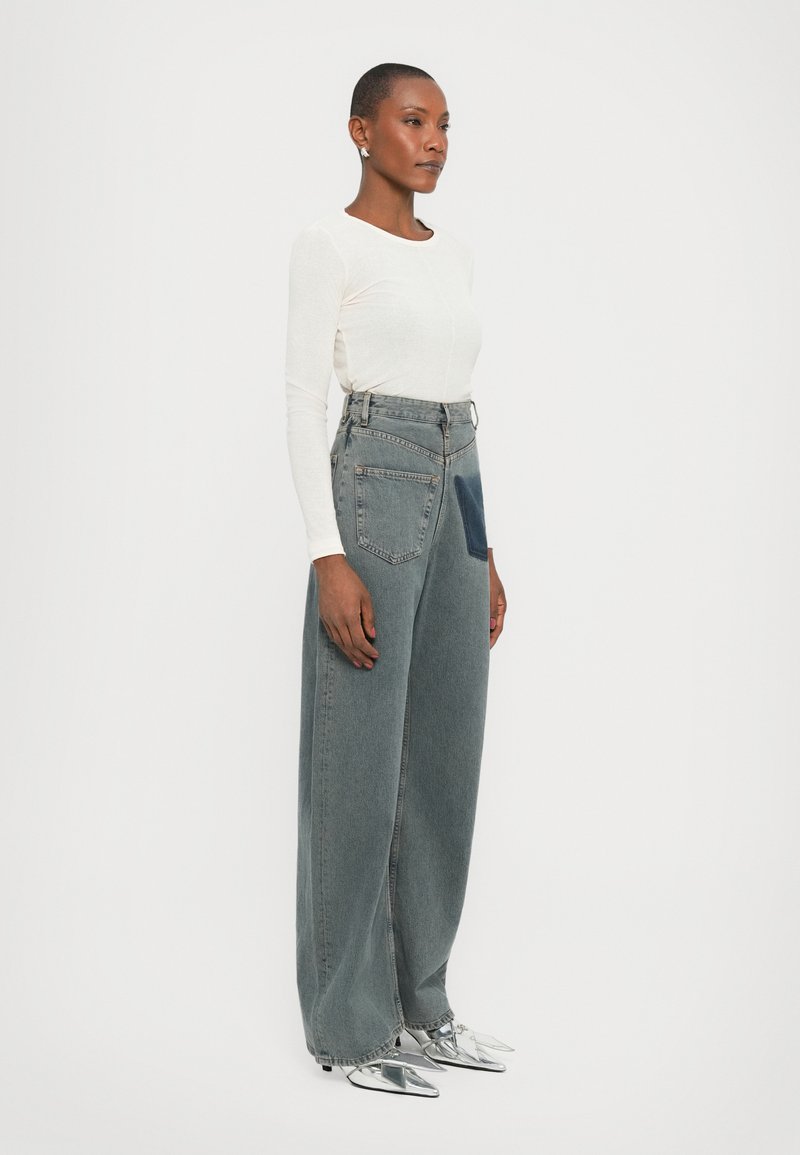 Jean taille haute à jambe large en denim gris clair, présentant une poche appliquée en tissu bleu foncé ; associé à un haut ajusté à manches longues blanc et des chaussures argentées.