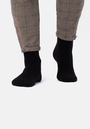 Beine in karierten Hosen mit Reißverschlüssen am Knöchel, hochgekrempelt, kombiniert mit einfarbigen schwarzen Socken vor weißem Hintergrund.