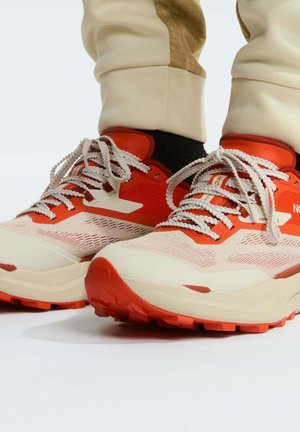 Beige en oranje wandelschoenen met dikke zolen en patroonveters, gedragen bij beige broeken en zwarte sokken tegen een witte achtergrond.