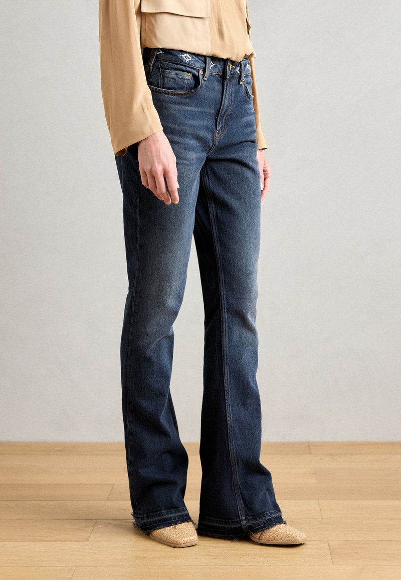 Mørkeblå denim flared jeans med fem lommer, lukking med knapp og glidelås, og subtil fading. Brukt med lyse teksturerte sko.