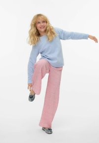 Pull bleu clair, pantalon large rayé rose et blanc, chaussures grises sans lacets avec un motif de rose, pose décontractée, fond blanc uni.