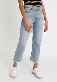 Jeans de mezclilla azul claro con corte recto, ligero desgaste en la rodilla y dobladillos deshilachados, combinados con zapatillas blancas.