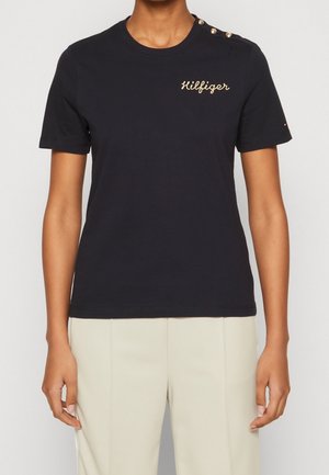Persoon die een zwart T-shirt met korte mouwen draagt met gouden "Hilfiger" tekst en knopen op de linkerschouder, gecombineerd met beige broek.