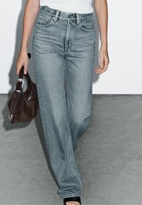 Jeans azzurri chiari con vita alta e vestibilità comoda, dotati di cinque tasche e texture sbiadita. Abbinati a una borsa marrone e sandali neri.