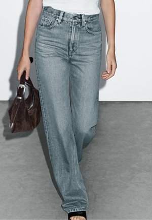 Lichtblauwe high-waisted jeans met een relaxte pasvorm, voorzien van vijf zakken en een vervaagde textuur. Gecombineerd met een bruine handtas en zwarte sandalen.