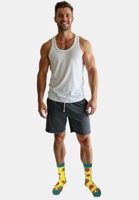 Hvid tanktop, grå shorts og gule sokker med røde jordbærmønstre. Atleten står selvsikkert og viser en muskuløs kropsbygning.