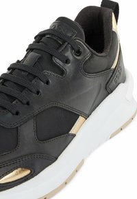 Baskets noires et dorées avec semelle épaisse blanche, col rembourré et lacets noirs, affichant un embossage de la marque sur la languette et le talon.
