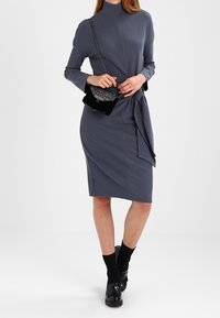 Abito grigio a maniche lunghe con scollo alto e vita con laccio. Abbinato a una piccola borsa nera e calze nere, indossato con scarpe nere.