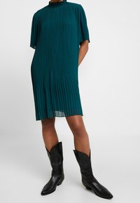 Teal-harmaatettu mekko, jossa on korkea kaulus ja lyhyet hihat, yhdistettynä terävään mustaan nahkasaappaaseen, jossa on monimutkaista ompelua.