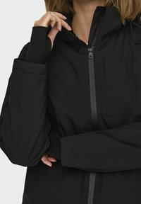 Veste noire légère en tissu lisse, dotée d'une fermeture éclair intégrale, d'une capuche ajustable, d'une poche zippée et de poignets avec orifices pour les pouces.