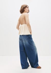 Kvinna i en ärmlös, krämbelagd stickad topp med öppen rygg och vida jeans i blå denim, stående med händerna i fickorna.