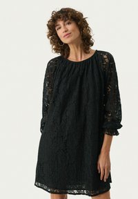 Schwarzes Spitzenkleid mit langen durchsichtigen Ärmeln, gerafftem Ausschnitt und lockerer Passform. Besitzt florale Muster und rüschengestaltete Ärmelenden.