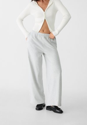 Woman wearing a white ribbed zip-up top, light gray wide-leg pants, and black loafers standing with hands in pockets.