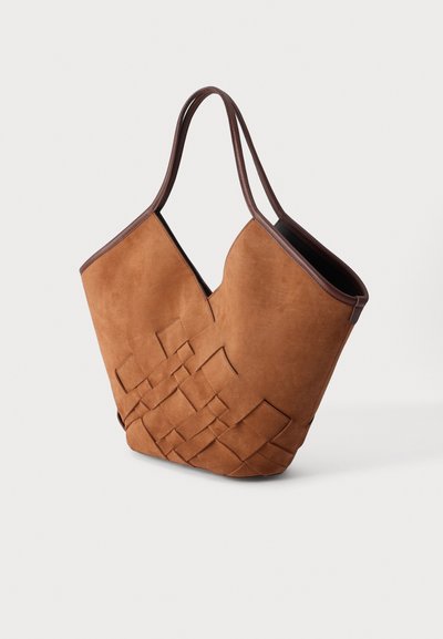 Sac cabas en daim marron avec panneau tissé et anses bordées de cuir marron foncé sur fond uni.