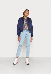 Casaco bomber azul-marinho sobre uma blusa com padrão floral, combinado com jeans de lavagem clara e rasgados e ténis brancos com detalhes em rosa.