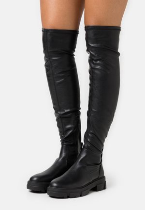 Černé kožené kozačky nad kolena s robustními podrážkami a bočními zipy, nošené na holých nohách na jednobarevném pozadí.