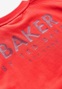 Sudadera roja con la inscripción "BAKER" en letras plateadas elevadas, con "BY TED BAKER EST. 1988" debajo en un tamaño de letra más pequeño. Material de algodón suave.