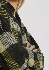 Černý a zelený kostkovaný pletený svetr s žlutými akcenty; silná textura, stojací límec, žebrované manžety a viditelné detaily šití.