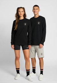 Svarta långärmade tröjor med en färgglad logotyp, kombinerade med svarta shorts och grå shorts. Båda bär svarta strumpor med regnbågsränder och vita sneakers.