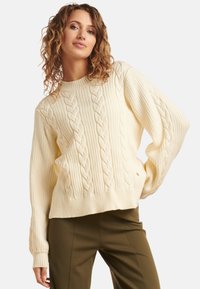 Cream cable-knit sweater with a ribbed texture, wide sleeves, and a round neckline. Paired with olive green pants.