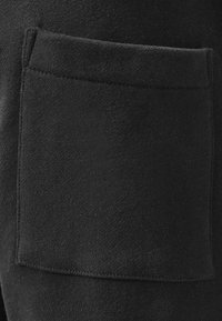 Tela de sudadera negra con un bolsillo cuadrado de superficie texturizada, ubicado en el lado derecho, cosido con hilo en contraste.