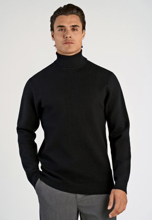 Zwarte coltrui van gladde stof, met een getailleerd ontwerp en geribbelde manchetten. Draagt met grijze broek.