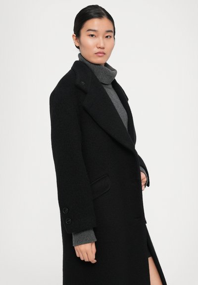 Femme portant un manteau en laine noir par-dessus un pull à col roulé gris, se tenant devant un fond blanc uni.