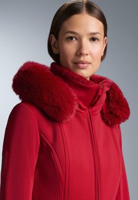 Piros külső ruha, texturált, cipzáras kialakítással és puha műszőrme gallérral. Az anyag simának tűnik, függőleges mintázattal a testen.