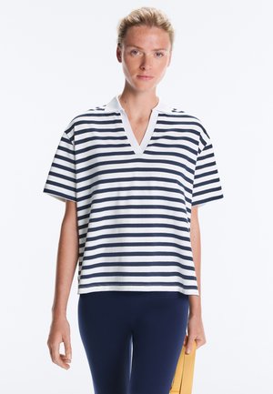 Femme portant un polo à manches courtes rayé horizontalement blanc et bleu marine et un pantalon bleu marine, tenant une sangle jaune dans sa main droite.