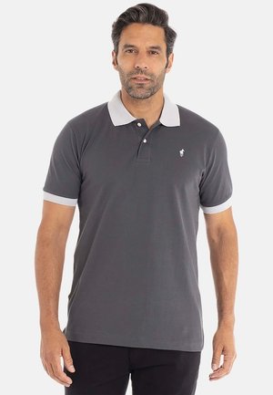 Homme portant un polo gris foncé avec un col blanc et des poignets blancs, se tenant devant un fond clair uni.