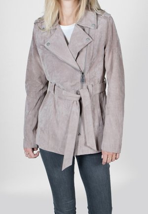 Kvinde iført en lys mauve ruskind jakke med bælte, asymmetrisk lynlås og mørke jeans, stående mod en ensfarvet baggrund.