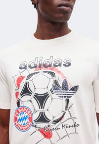 Άνδρας που φοράει λευκό μπλουζάκι Adidas με γραφικό ποδοσφαιρικής μπάλας, λογότυπο FC Bayern München και κείμενο Bayern München σε μαύρο και κόκκινο.