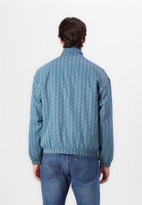 Light blue jacket with a geometric pattern, elastic cuffs and waist, and a stand-up collar. Paired with denim jeans.