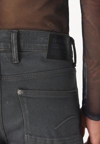 Szürke farmernadrág texturált felülettel, fekete bőr címkével, standard zsebekkel és fém gomb részletekkel.