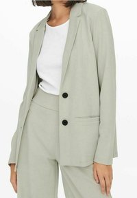 Lichtgrijze blazer met een gestructureerd ontwerp, sluiting met één knoop en een borstzak. Ggedragen over een wit T-shirt. Gladde textuur.