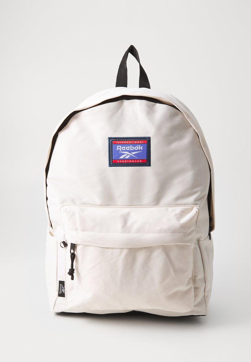 Mochila de lona beige con un asa negra, bolsillo frontal con cremallera y logo de Reebok bordado en un parche azul y rojo. Presenta una superficie texturizada.