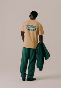 T-shirt beige à manches courtes avec un graphique floral au dos, associé à un pantalon de survêtement vert foncé et une veste assortie, porté avec des chaussures noires.