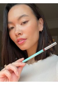 En myntgrön läppennna med en silvertip hålls bredvid en person med rakt svart hår iförd en ribbad vit topp.