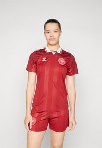 Piros foci mez csíkozott mintával, rövid ujjú, fehér gallérral, és sportlogóval a mellkason és a nadrágon. Könnyű anyagból készült.