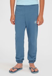 Blå bomullsjoggingbyxor med elastiska cuffar, med en sidoficka och en vit solgrafiklogotyp på vänster lår. Bärs med flip-flops.